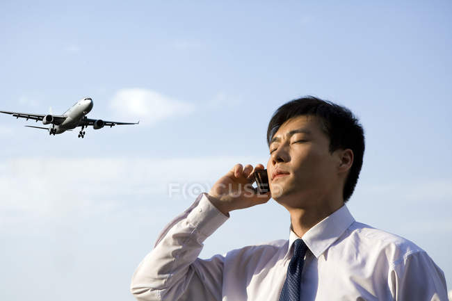 Chinese businessman talking on phone in front of sky with airplane — Stock Photo