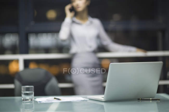 Ordinateur portable sur table de bureau avec femme d'affaires parlant au téléphone en arrière-plan — Photo de stock