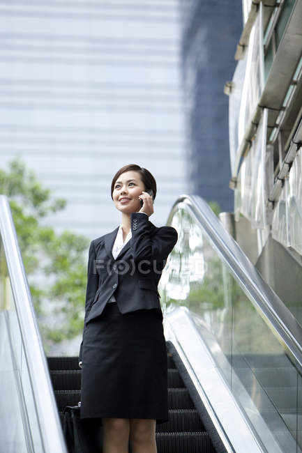 Chinese businesswoman standing on escalator and talking on phone — Stock Photo