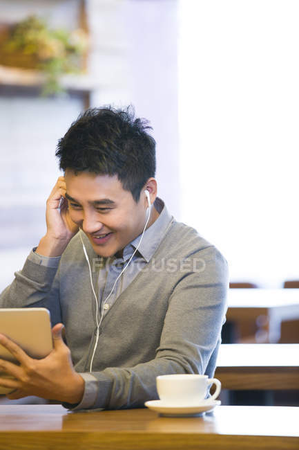 Chinese man using digital tablet with earbuds in coffee shop — Stock Photo