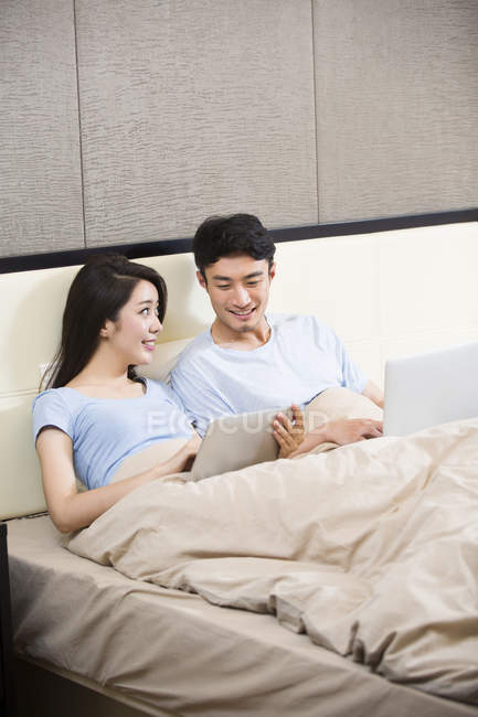 Young Chinese couple using wireless devices in bed — Stock Photo
