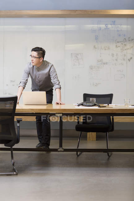 Chinese man standing with laptop and thinking in board room — Stock Photo