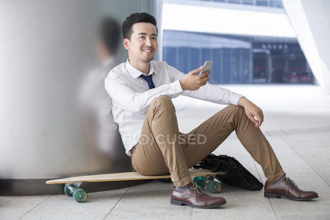 Uomo d'affari cinese seduto su skateboard con smartphone — Foto stock