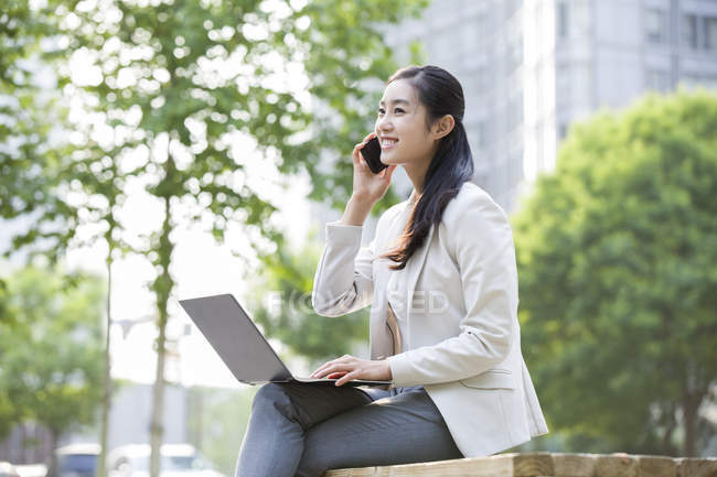 Empresaria sentada con laptop y hablando por teléfono - foto de stock