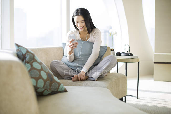 Mujer china escuchando música en el sofá en el interior del hogar - foto de stock
