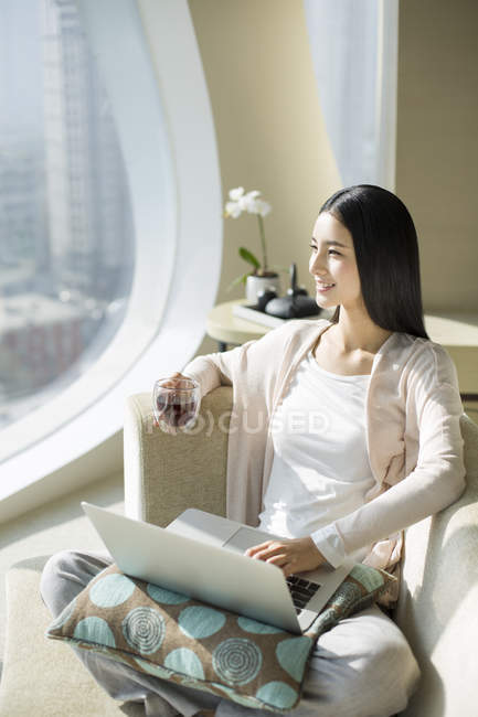 Femme chinoise avec tasse à thé en utilisant un ordinateur portable sur le canapé — Photo de stock