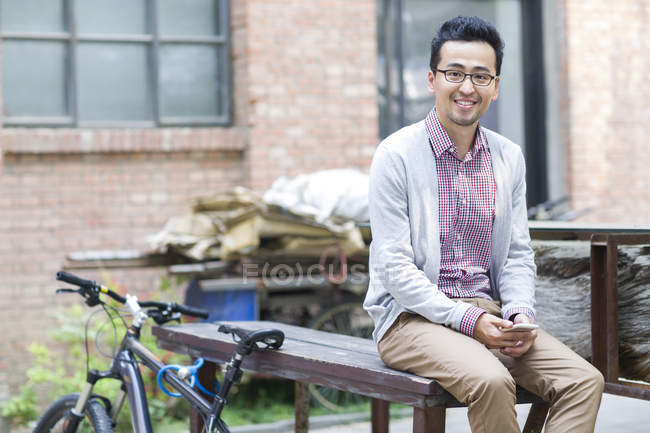 Hombre chino sentado en el banco con teléfono inteligente y bicicleta - foto de stock