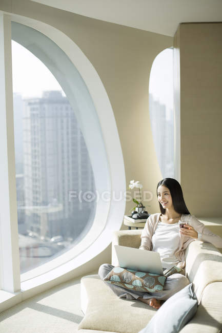 Chinese woman sitting with tea cup and laptop on couch — Stock Photo