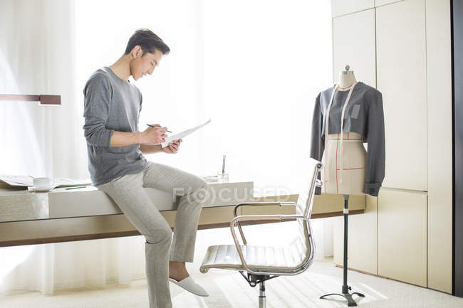 Diseñador de moda chino sentado en la mesa y mirando bocetos - foto de stock