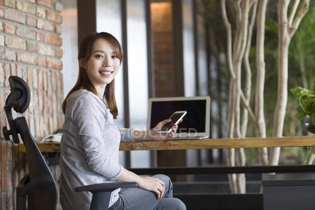 Chinese woman holding smartphone at office desk — Stock Photo