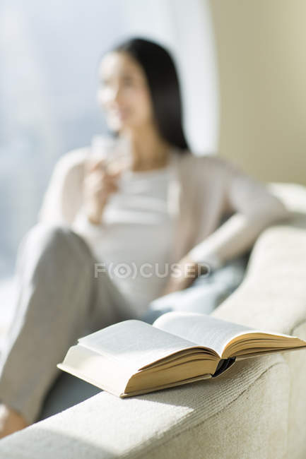 Primer plano del libro abierto con la mujer sentada en el sofá en el fondo - foto de stock