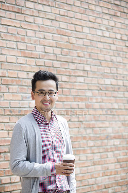 Chinese man holding cup of coffee in front of brick wall — Stock Photo