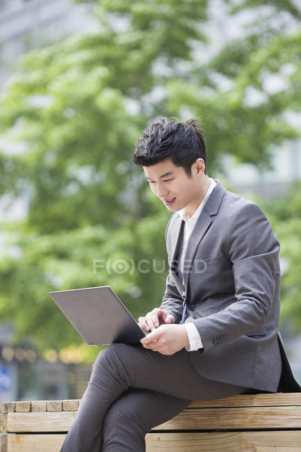 Uomo d'affari cinese che lavora con il computer portatile sulla panchina stradale — Foto stock