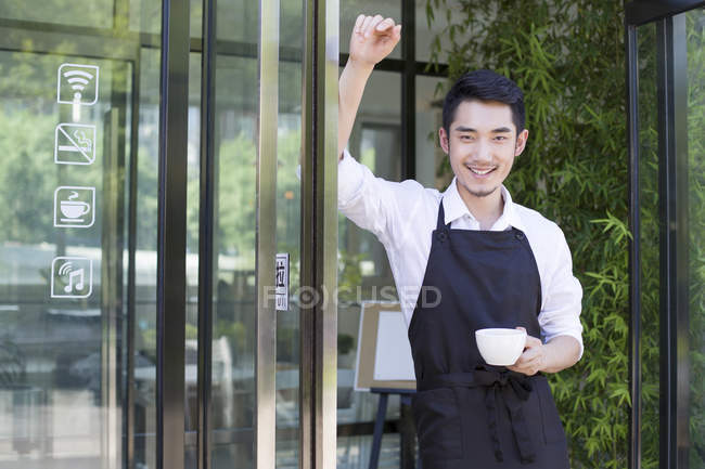 Shopkeeper standing in doorway of cafe with cup of coffee — Stock Photo