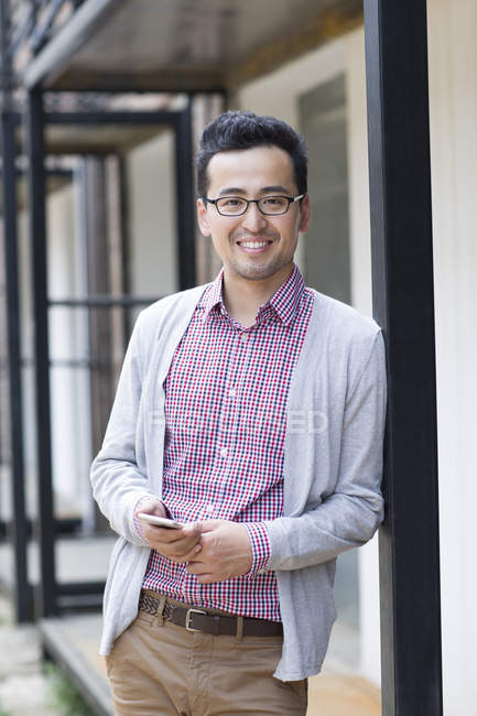 Hombre chino sosteniendo el teléfono inteligente y apoyado en la pared - foto de stock