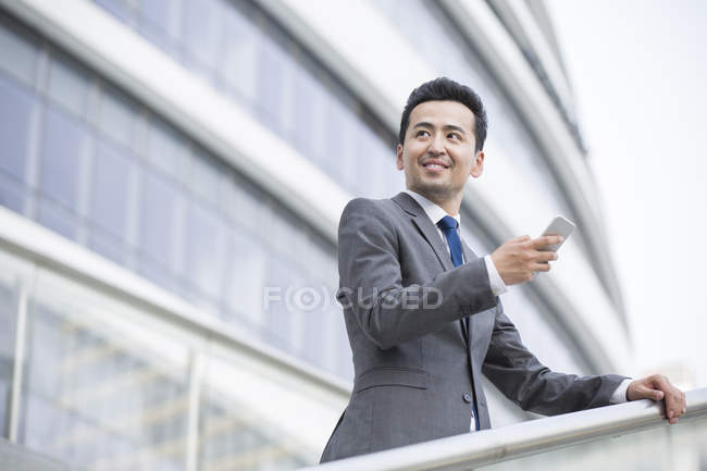 Chinese businessman standing in front of office building with smartphone — Stock Photo