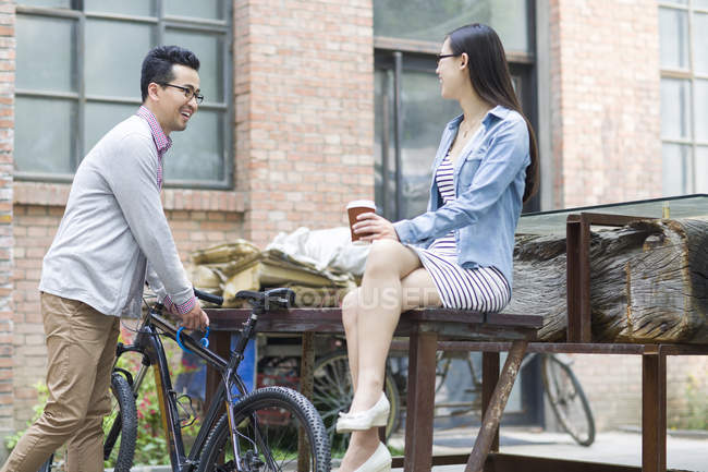 Mujer china sentada en el banco con café y hablando con el hombre con la bicicleta - foto de stock