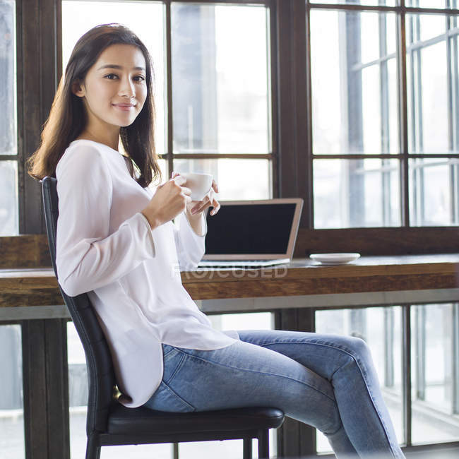 Chinese woman drinking coffee in cafe with laptop on table — Stock Photo