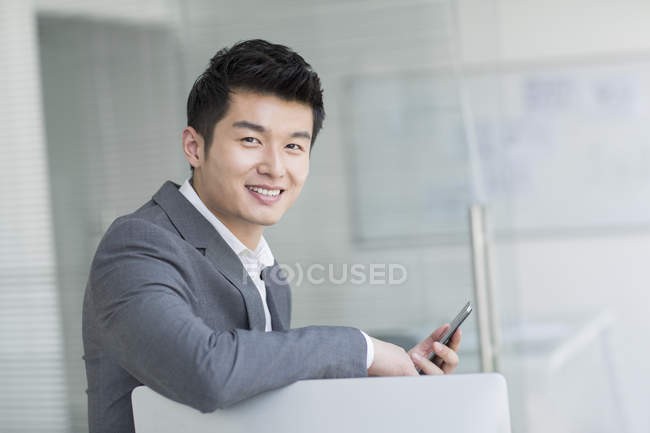 Hombre de negocios chino sentado con teléfono inteligente en la oficina - foto de stock