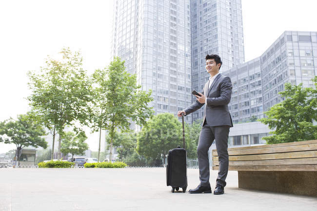 Chinese businessman holding suitcase and smartphone on street — Stock Photo