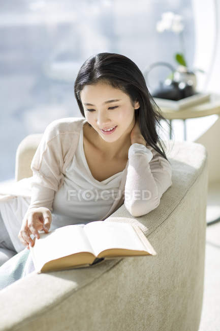 Mujer china leyendo libro en sofá en el interior del hogar - foto de stock