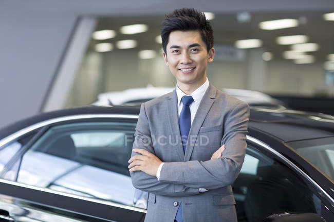 Chinese salesman standing in car showroom with arms folded — Stock Photo