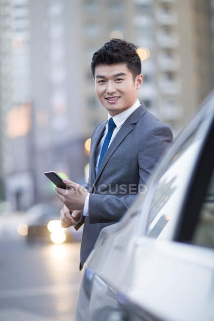 Chinese businessman standing at car with smartphone — Stock Photo