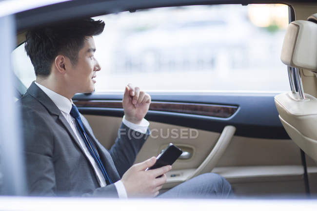 Homme d'affaires chinois assis sur le siège arrière de la voiture avec smartphone — Photo de stock