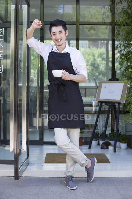 Shopkeeper de pie en la puerta de la cafetería con taza de café - foto de stock