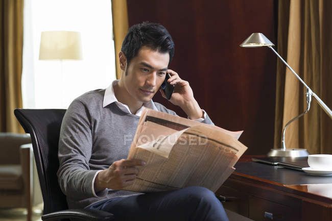 Chinese man talking on phone with newspaper in home interior — Stock Photo