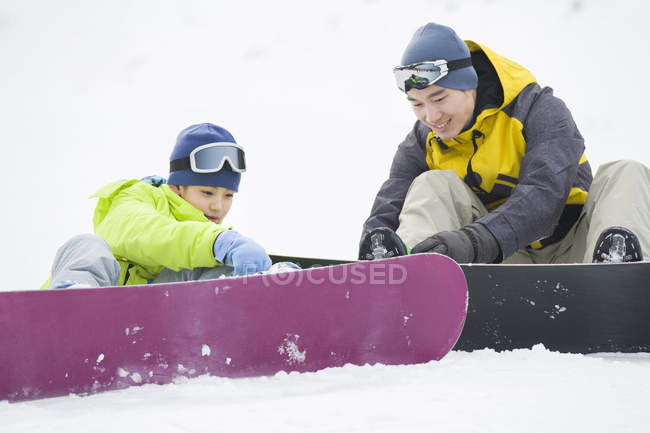 Padre chino ayudando a su hijo con equipo de snowboard en la nieve - foto de stock