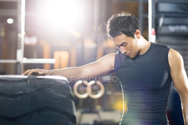 Китаец, опирающийся на большую шину в тренажерном зале и смотрящий вниз — стоковое фото