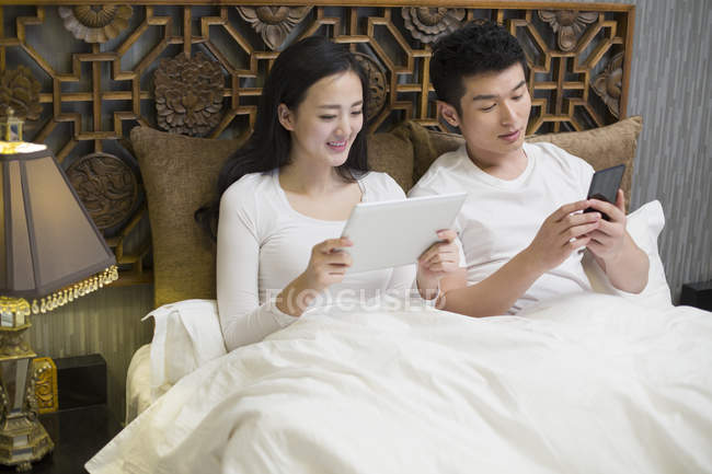 Casal chinês usando tablet digital e smartphone na cama — Fotografia de Stock