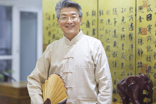 Sênior chinês homem segurando vintage ventilador portátil no interior tradicional — Fotografia de Stock