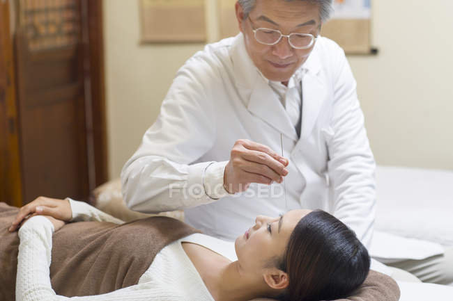 Médico senior que realiza tratamiento de acupuntura en la cara femenina - foto de stock
