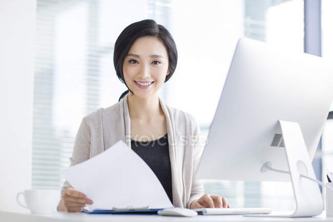Mujer china sentada con papeles en la oficina y mirando en cámara - foto de stock