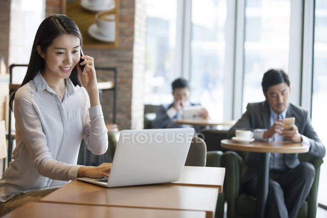 Chinese woman talking on phone and using laptop in cafe — Stock Photo