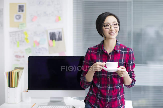 Femme chinoise tenant une tasse de café et regardant loin dans le bureau — Photo de stock