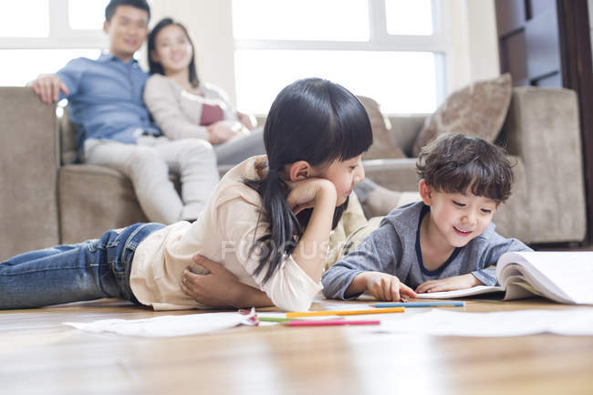 Irmãos chineses estudando juntos no chão com os pais no sofá assistindo — Fotografia de Stock