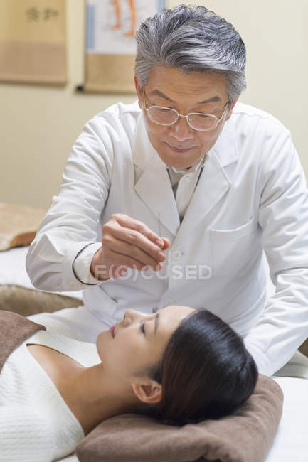 Médico senior que realiza tratamiento de acupuntura en la cara femenina - foto de stock