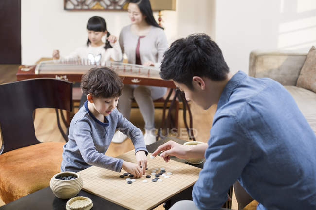 Padre e hijo jugando juego de Go con la madre y la hija tocando el instrumento musical en segundo plano - foto de stock