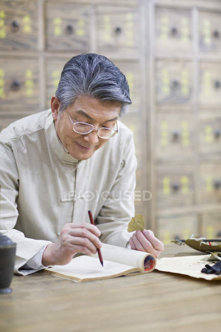 Médico chino mayor sosteniendo hierba medicinal y escribiendo en cuaderno - foto de stock
