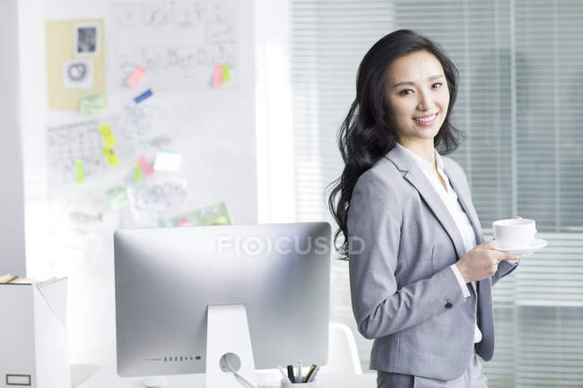 Chinesische Geschäftsfrau hält eine Tasse Kaffee in der Hand und blickt in die Kamera am Arbeitsplatz — Stockfoto