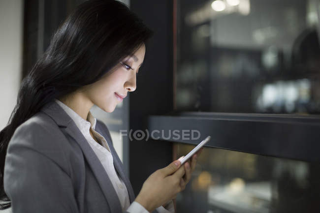 Empresária chinesa usando smartphone pela janela do escritório — Fotografia de Stock