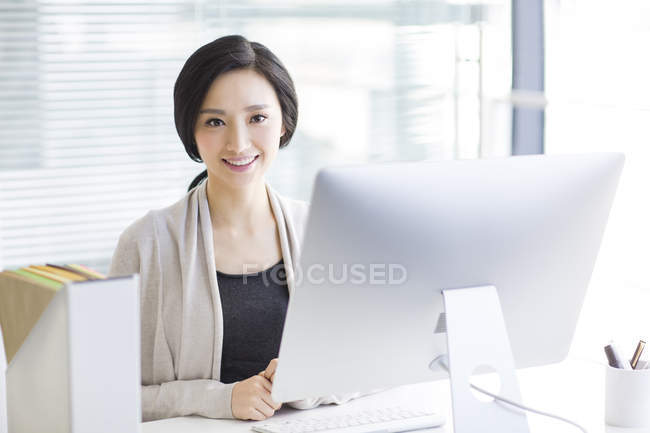 Mujer china sentada en el escritorio en la oficina y mirando en la cámara - foto de stock