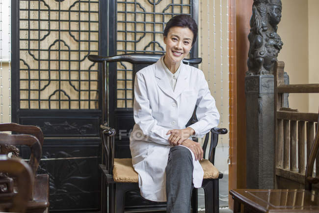 Mujer china doctor sentado en silla y mirando en cámara - foto de stock
