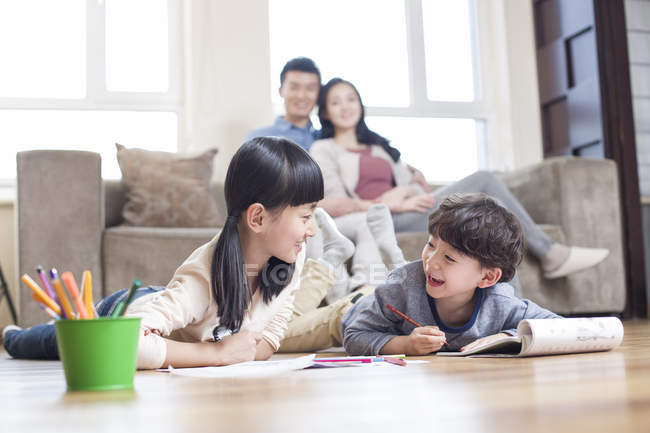 Irmãos chineses estudando juntos no chão com os pais no sofá assistindo — Fotografia de Stock