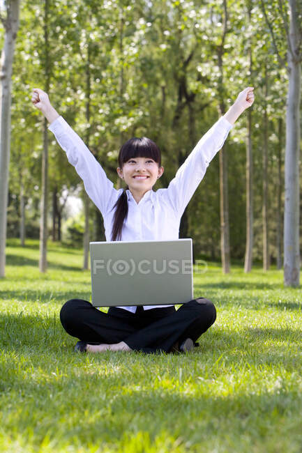 Mujer china sentada en la hierba en el parque con portátil y levantando las manos - foto de stock