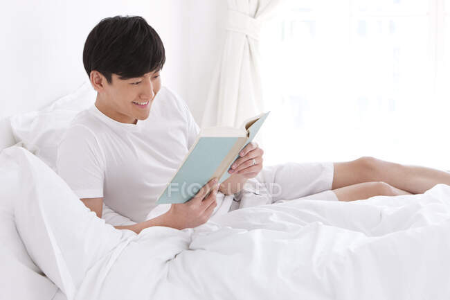 Joven chino leyendo un libro en la cama - foto de stock