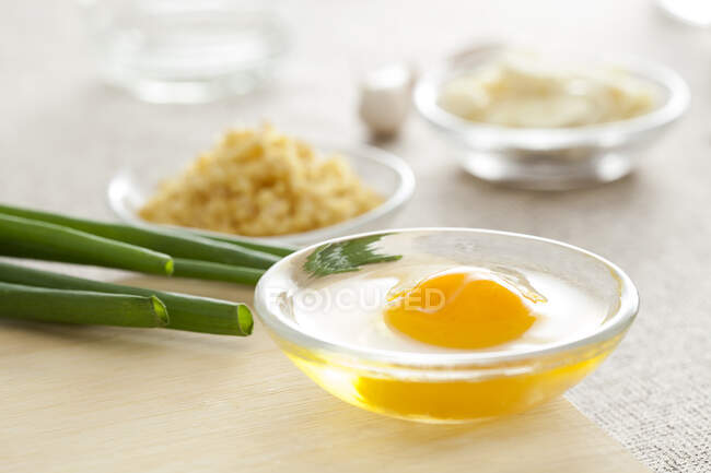 Huevo en tazón de vidrio con cebolla verde en la superficie de madera con otros ingredientes desenfocados - foto de stock